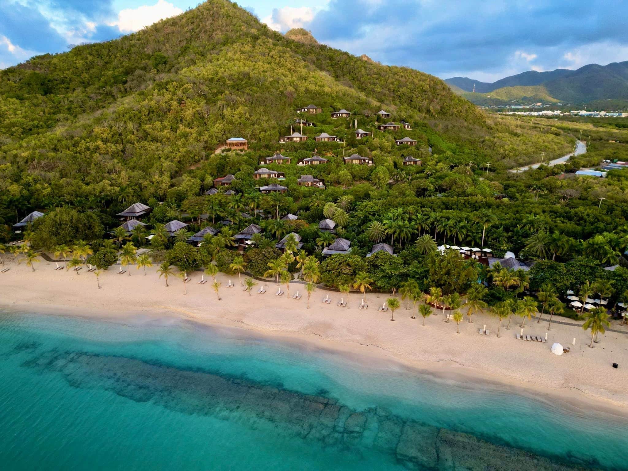 Aerial view of Hermitage Bay Antigua showing the beachfront, turquoise Caribbean waters, tropical landscaping, and hillside villas overlooking the resort.