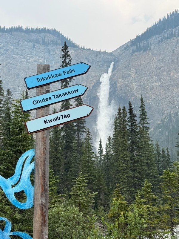 Directional sign for Takakkaw Falls with the waterfall and mountains visible in the background