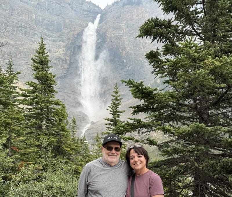 Couple standing in front of Takakkaw Falls surrounded by evergreen trees in Yoho National Park